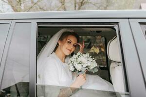 une magnifique souriant la mariée sur sa mariage journée est séance dans une voiture et en portant une mariage bouquet de blanc et rose des roses. une Jeune femme avec une magnifique coiffure se réjouit sur sa mariage journée photo