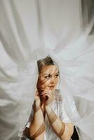 fermer coup de un élégant brunette la mariée dans une blanc robe posant en dessous de une voile fermer. la mariée portrait, professionnel mariage maquillage et coiffure, de mariée mode. magnifique la mariée dans une voile photo