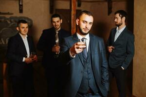 le jeune marié dans le premier plan et le sien copains dans élégant costume en buvant whisky dans le Hôtel chambre, le Matin avant le mariage les préparatifs photo