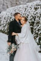 sensible portrait de content jeunes mariés. le jeune marié câlins et baisers le la mariée dans le hiver parc. le la mariée dans une mariage robe et une blanc poncho. le jeune marié est habillé dans une noir manteau. photo