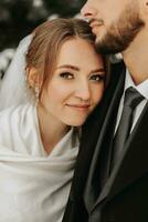 portrait de le la mariée embrassé par le jeune marié. jeune marié et la mariée dans le hiver parc. le la mariée dans une mariage robe et poncho. jeune marié dans une noir manteau. photo