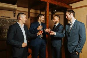 le jeune marié et le sien copains dans élégant costume boisson whisky dans le Hôtel chambre, le Matin avant le mariage les préparatifs. groupe de Hommes parlant et en buvant whisky photo