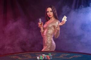 fille en jouant poker à casino, en portant verre de Champagne, deux as. posant à table avec puces, argent sur il. noir, fumée Contexte. fermer. photo