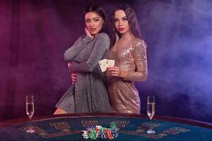 deux les filles sont en jouant poker à casino, étreindre, en portant cartes, posant à une table avec puces, Champagne sur il. noir, fumée Contexte. fermer. photo