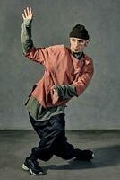 gracieux homme avec tatoué corps, des boucles d'oreilles, barbe. habillé dans chapeau, décontractée vêtements et noir baskets. dansant sur gris Contexte. salle de danse, hip hop photo