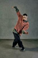 majestueux compagnon avec tatoué affronter, barbe. habillé dans coloré sauteur, noir un pantalon et baskets. dansant, gris Contexte. salle de danse, hip hop photo