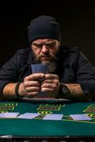 sérieux barbu homme séance à poker table et en portant cartes isolé sur noir photo