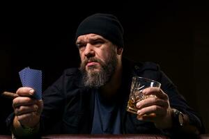 barbu homme avec verre de de l'alcool boisson et cartes dans mains séance à poker table sur noir Contexte photo