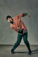 fort Masculin avec tatoué affronter, barbe. habillé dans chapeau, coloré sauteur, vert pantalon, noir baskets. dansant sur gris Contexte. salle de danse, hip hop photo