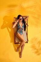 magnifique Jeune brunette femme dans coloré maillot de bain mensonges sur le fainéant sur Jaune Contexte photo