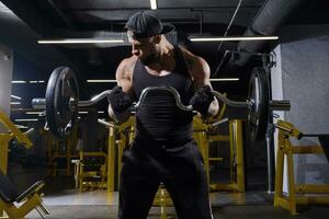 tatoué Masculin dans noir sport gants, shorts, gilet et casquette. levage haltère, formation le sien biceps, permanent dans foncé Gym avec Jaune équipement. proche en haut photo