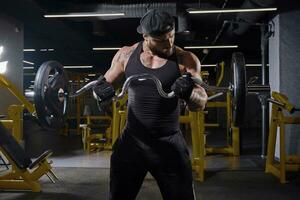 barbu homme dans noir sport gants, shorts, gilet et casquette. levage une haltère, formation le sien muscles, permanent dans foncé Gym avec Jaune équipement. proche en haut photo