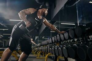 tatoué, musclé gars dans noir shorts, gilet et casquette. exercice avec haltères pour formation le sien triceps dans foncé salle de sport. sport, aptitude. proche en haut photo