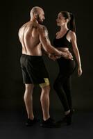 athlétique homme dans short et baskets avec brunette femme dans leggings et Haut posant sur noir Contexte. aptitude couple, Gym concept. photo