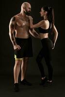 athlétique homme dans short et baskets avec brunette femme dans leggings et Haut posant sur noir Contexte. aptitude couple, Gym concept. photo