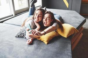 deux soeurs jumelles souriantes mignonnes tenant un smartphone et faisant un selfie. les filles s'allongent sur le canapé posant et joie photo