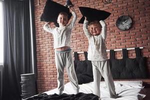 vilains enfants petit garçon et fille ont organisé une bataille d'oreillers sur le lit dans la chambre. ils aiment ce genre de jeu photo