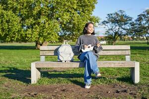 insouciant fille est assis sur banc dans parc avec ukulélé, pièces et chante en plein air sur ensoleillé content journée photo