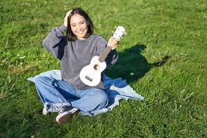 excité asiatique fille est assis dans parc avec ukulélé, pièces instrument et se sent content optimiste, positif des moments et gens concept photo