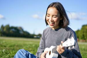 content gens et loisirs. souriant asiatique fille en jouant ukulélé guitare et en chantant, séance dans parc en plein air sur couverture photo