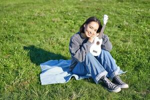 mignonne romantique asiatique fille séance avec sa instrument dans parc, étreindre ukulélé et souriant, repos en plein air photo