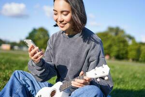 La technologie et musique. souriant asiatique branché fille séance dans parc avec téléphone intelligent et en portant ukulélé, en jouant instrument photo