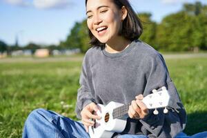 portrait de magnifique asiatique femme en chantant, en jouant ukulélé guitare dans parc, séance seul sur herbe sur ensoleillé journée photo