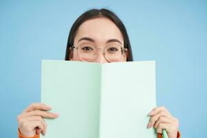 portrait de mignonne Japonais femme dans lunettes, furtivement de carnet de notes, couvertures visage avec sa Remarques, sourit avec yeux, bleu Contexte photo