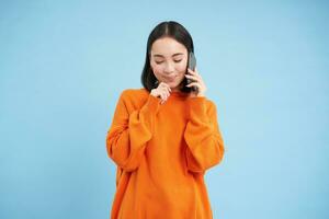 Jeune femme avec réfléchi regarder, réponses téléphone appel, pourparlers sur mobile et souriant, en pensant avec heureux affronter, permanent plus de bleu Contexte photo