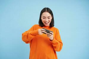 content souriant fille, 25 années, pièces mobile vidéo Jeux, détient téléphone intelligent dans tous les deux mains, des stands contre bleu Contexte photo