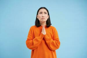 prier à Dieu. misérable asiatique fille détient mains dans supplication, fait du souhait, regards en haut et attend pour miracle, des stands plus de bleu Contexte photo