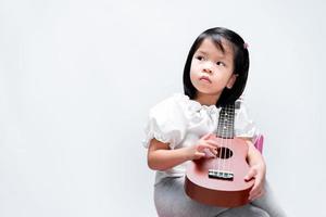 adorable petite fille met son ukulélé sur son épaule. enfant joue des instruments de musique, regardant de côté. sur fond blanc de studio. photo