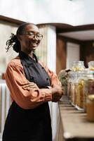 détaillé image de un africain américain femelle commerçant avec un tablier souriant à le caméra près verre bocaux. portrait coup de une noir femme portant lunettes avec sa bras franchi plus de sa poitrine. photo