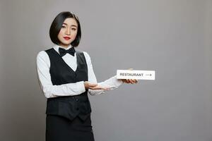 asiatique serveuse en portant restaurant direction signalisation et à la recherche à caméra tandis que posant dans studio. hospitalité un service femme employé dans uniforme montrant cafétéria aiguille acier signe portrait photo