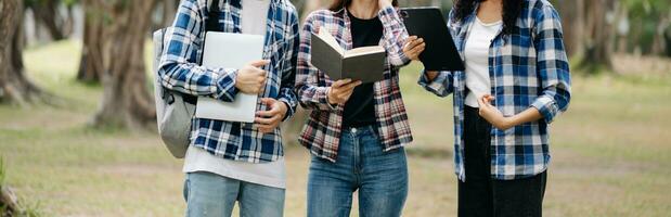 Trois Jeune Université élèves est en train de lire une livre tandis que relaxant séance sur herbe dans une Campus parc avec sa amis. éducation concept photo