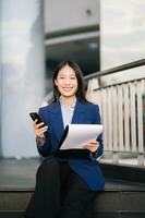 sur de soi asiatique femme avec une sourire permanent en portant bloc-notes et tablette à en dehors côté bureau. gros ville photo