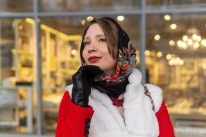 content rêveur Jeune femme dans une foulard dans du froid temps des stands à l'extérieur contre le Contexte de une floue brillant magasin fenêtre photo