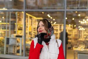 content rêveur Jeune femme dans une foulard dans du froid temps des stands à l'extérieur contre le Contexte de une floue brillant magasin fenêtre photo