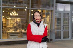 Jeune femme dans une foulard dans du froid temps des stands à l'extérieur contre le Contexte de une floue brillant magasin fenêtre photo