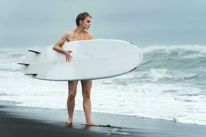 sexualité femme surfeur en marchant sur sablonneux plage avec blanc planche de surf contre Contexte océan vagues photo