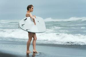 surfeur femme permanent sur sablonneux plage et porter blanc planche de surf contre Contexte de mer vagues photo