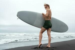 femelle surfeur permanent sur noir sablonneux plage en portant blanc planche de surf contre Contexte de mer vagues photo