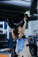 chevronné mécanicien travail sur suspendu voiture dans garage, vérification moteur pendant routine entretien. auto réparation magasin professionnel sous véhicule, inspecter Composants en utilisant travail lumière photo