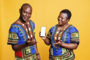 souriant noir couple portant ethnique se ferme montrant téléphone intelligent avec blanc Vide écran maquette. de bonne humeur homme et femme en présentant mobile téléphone avec vide écran tactile pour app La publicité photo