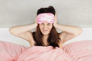 une magnifique fille est assis dans lit dans pyjamas et une sommeil masque. femme ne peux pas dormir, le voisins sont bruyant ou elle avoir mal de crâne rose lit lin photo