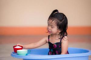 Jolie fille de 3 ans assise dans une douche en plastique bleu. enfant heureux humide d'utiliser un verre pour transférer l'eau. les enfants asiatiques ont un doux sourire. activités sympas. pendant l'été ou le printemps.