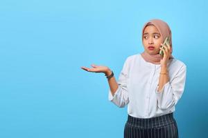 Portrait de triste jeune femme asiatique parlant au téléphone avec les mains levées sur fond bleu photo