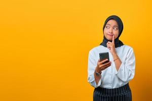portrait de jeune femme asiatique pensive tenant un téléphone portable et la main sur les lèvres sur fond jaune photo