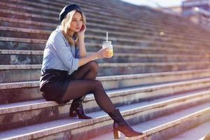 femme blonde avec béret, buvant un jus d'orange naturel dans un verre de cristal, assise sur quelques marches. photo