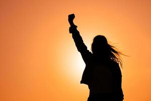 ai généré silhouette de une femme avec écoulement cheveux avec sa poing élevé en haut dans le le coucher du soleil ciel, fille Puissance photo